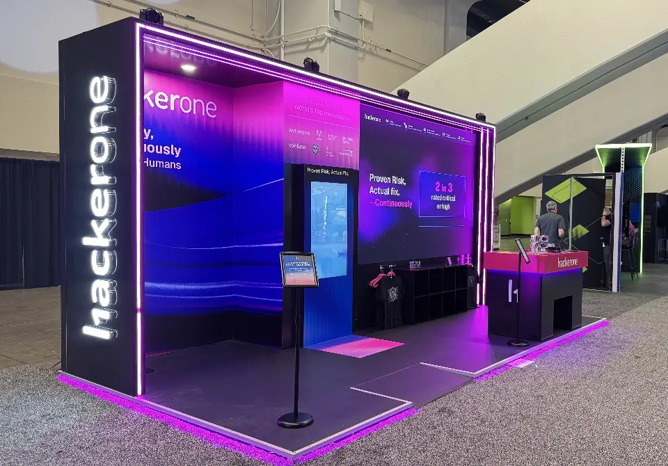 Angled view of the HackerOne stand showing the lit vertical logo column, kiosk and T-shirt shelving