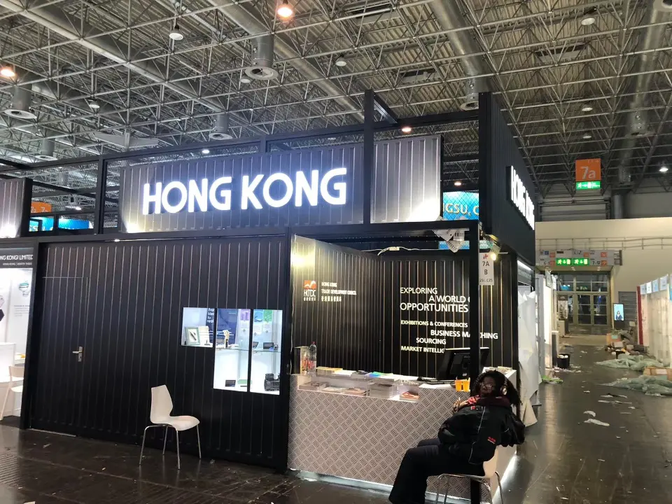 Black corner stand under HONG KONG lettering with an info counter, wall screens and a chair
