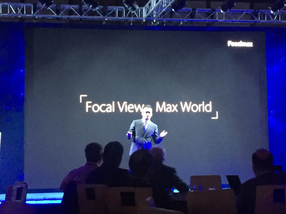 Presenter gesturing on stage with the slogan screen and blue edge lighting