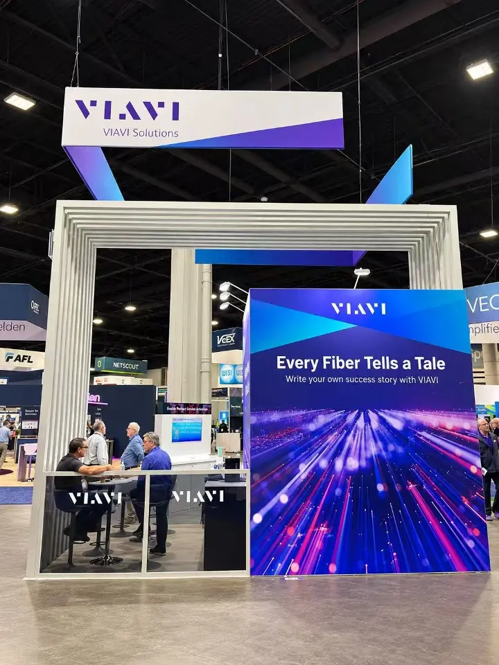 VIAVI Solutions meeting area behind a glass rail with VIAVI logos, beside a fiber-light graphic tower