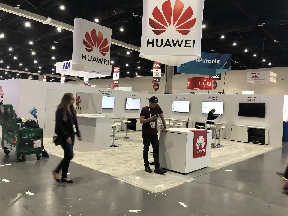 During build-up: white Huawei booth with tall hanging logo signs and a row of wall screens