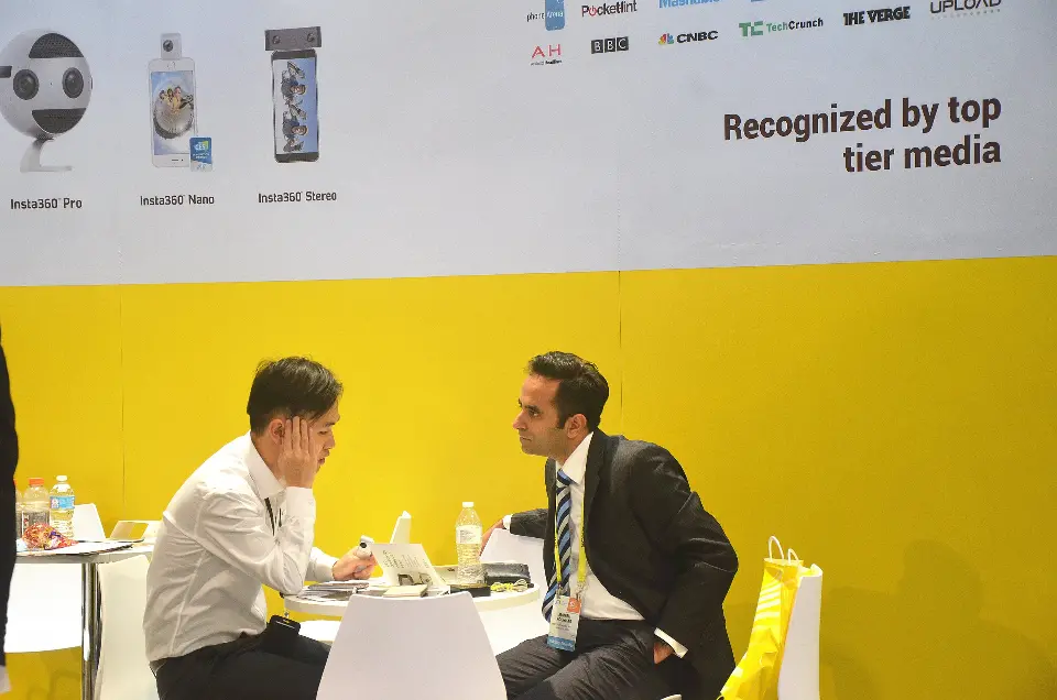 Two people talking at a white table beside a media logo wall