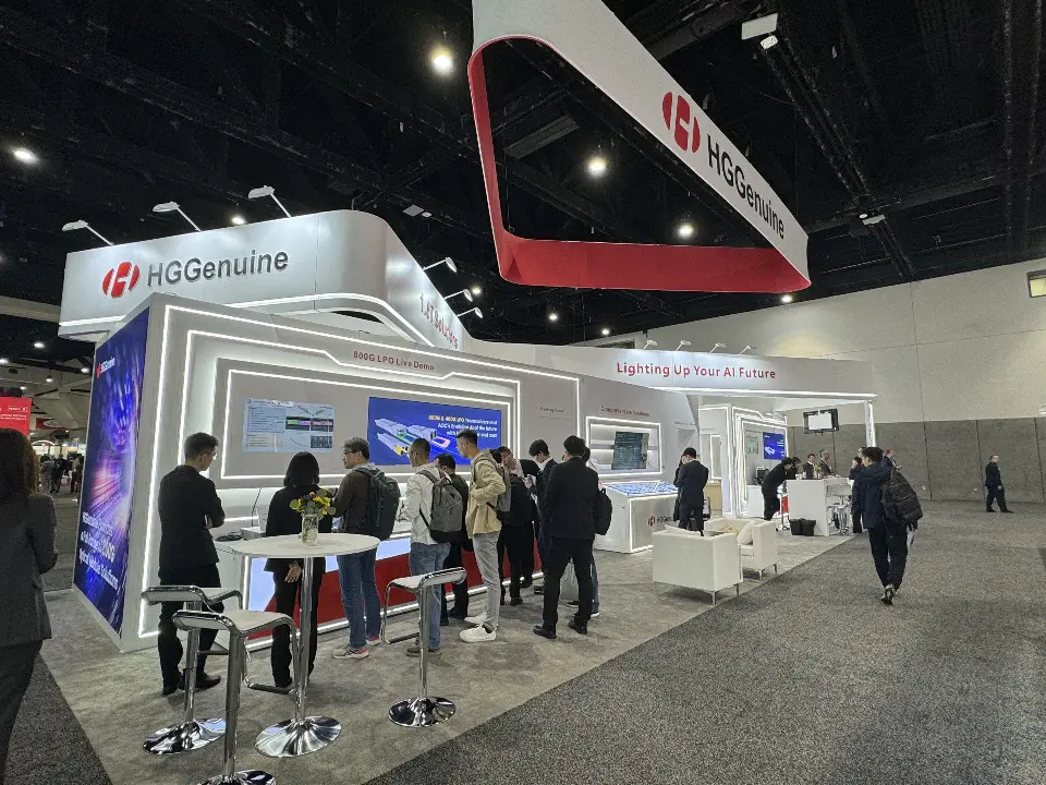 Front corner of the HGGenuine booth with high-top tables, visitors and red hanging header