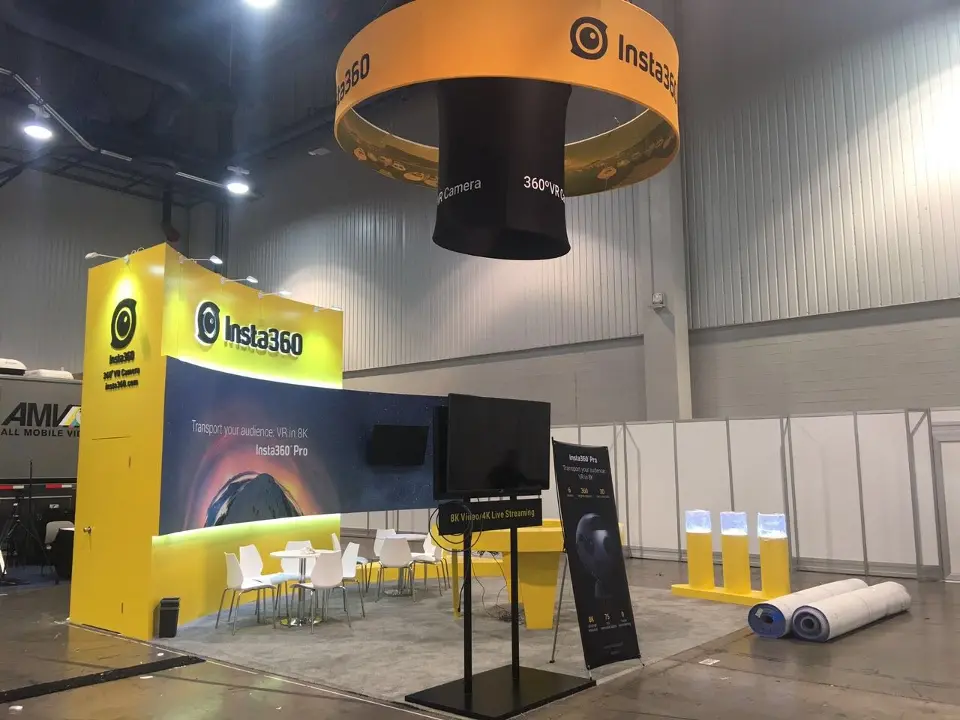 Insta360 booth corner with yellow wall, logo panel, white seating and a screen on a stand