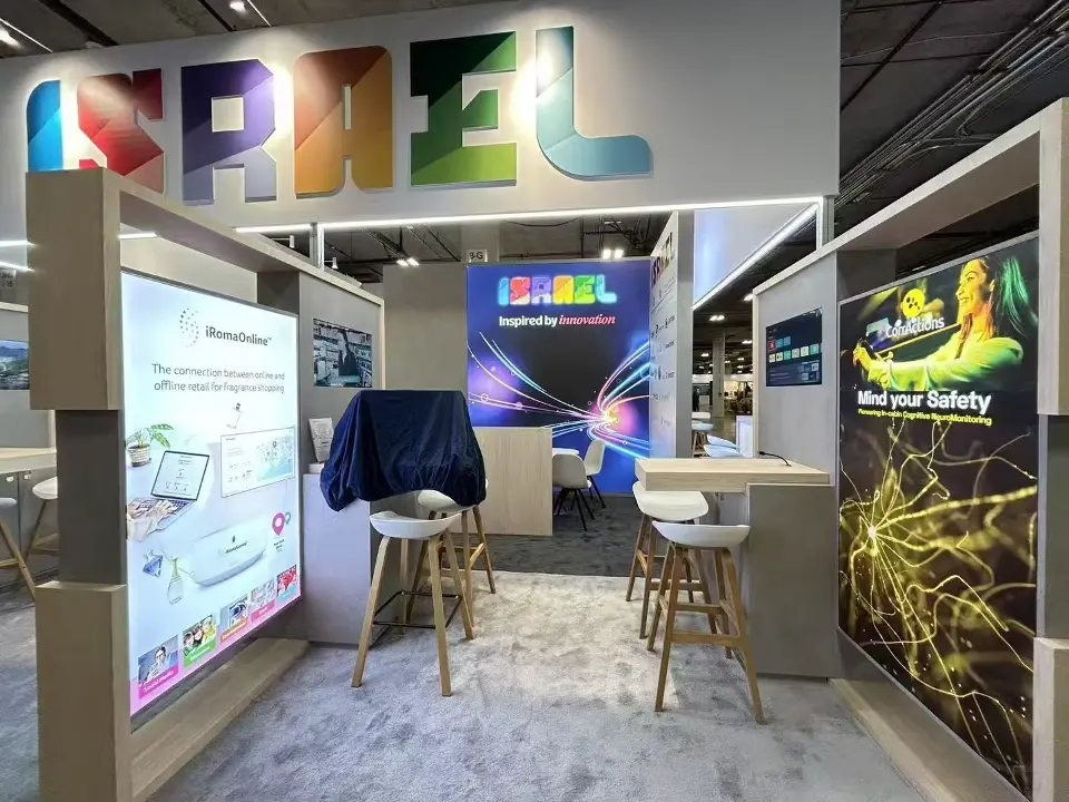 Exhibitor pod in the Israel Pavilion with backlit product graphics, stools and the colourful header behind