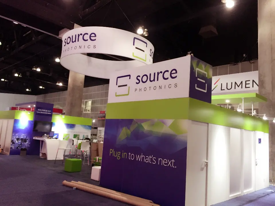 Corner of Source Photonics booth with purple tagline wall, green fascia and ring sign