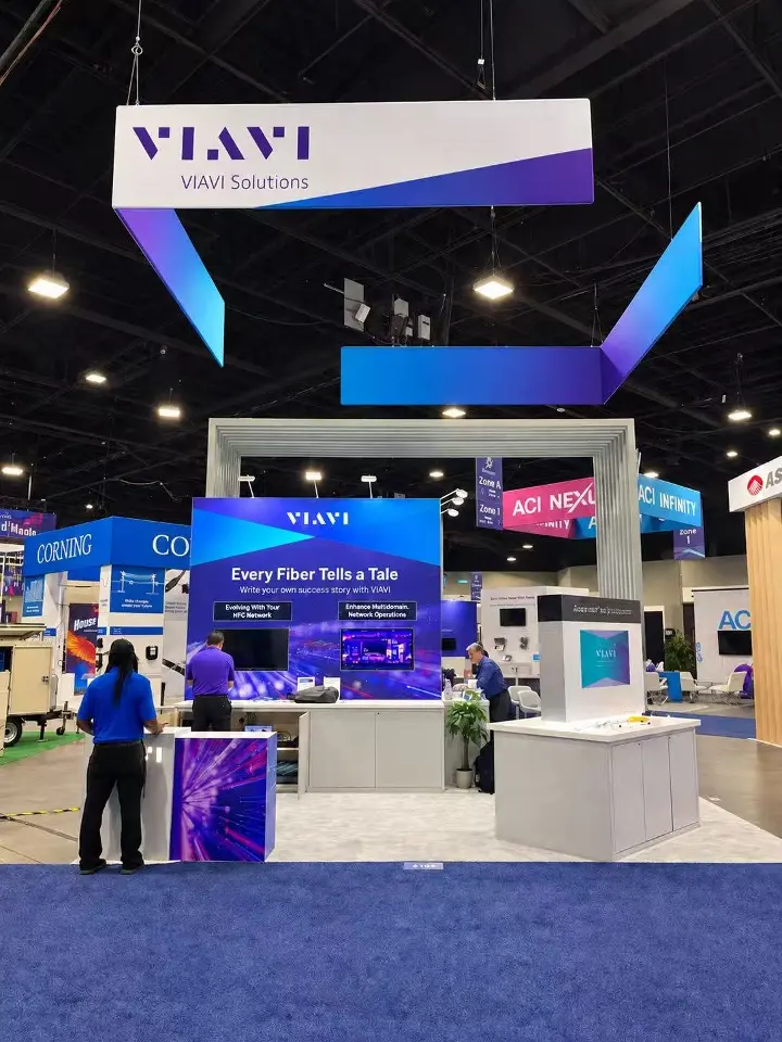 Front of the VIAVI Solutions booth with angled purple and blue hanging banners above a two-screen demo wall