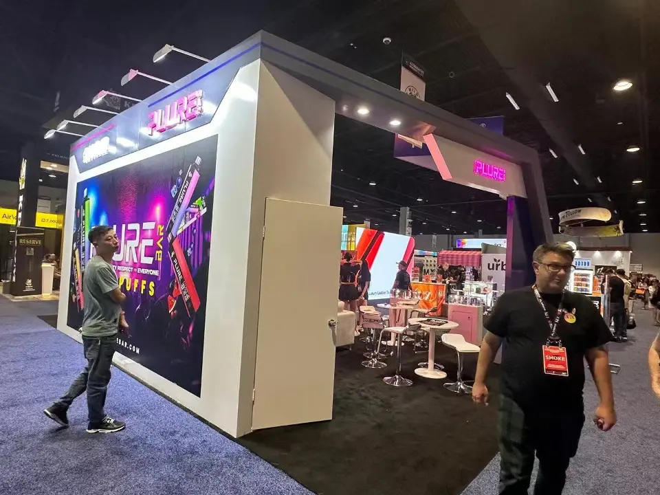Second angle of the backlit PLURE tower with visitors walking past the booth