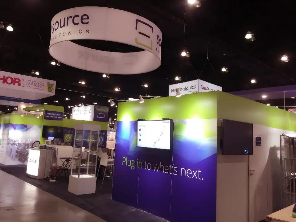 Hanging source photonics ring sign above purple walls and a wall-mounted screen