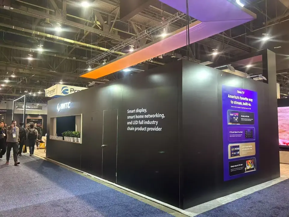 Long black side wall of the AMTC booth with product graphics under an orange overhead light band