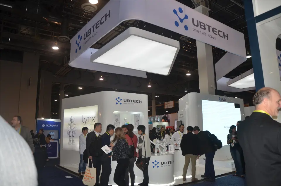 UBTECH booth with white hanging sign, lit ceiling panel and visitors at the counters