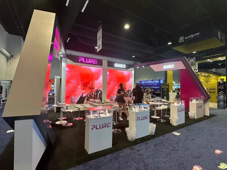 Corner of the PLURE booth with a tall white angled panel and red smoke graphic