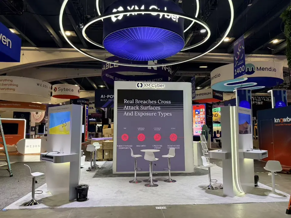 XM Cyber stand front with Real Breaches Cross Attack Surfaces wall, demo kiosks and white stools