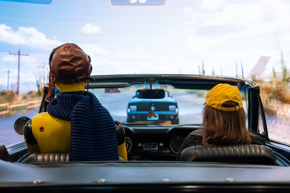 Figure in an aviator cap and a guest in the convertible watching the desert road on the LED wall